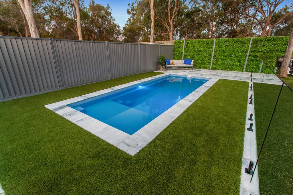 A modern, rectangular swimming pool surrounded by artificial grass and sleek white stone tiles. Green hedges are visible in the background, offering privacy to the pool area, which features a blue lounge chair beside the water.