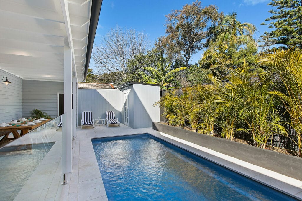 A backyard with a swimming pool surrounded by palm trees and greenery, featuring a patio area with lounge chairs. The house exterior shows light-colored walls and a wooden table set for dining.