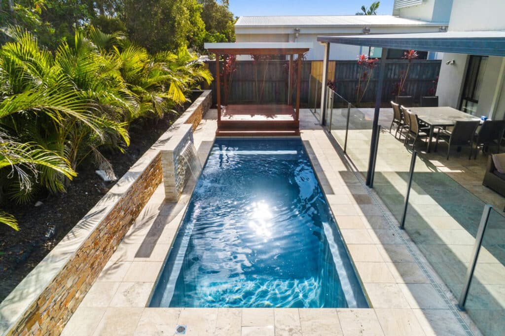 An overhead view of a modern swimming pool surrounded by palm trees and a stone wall. A wooden pergola with a seating area is near the pool, and a dining set is visible on a patio in the background.
