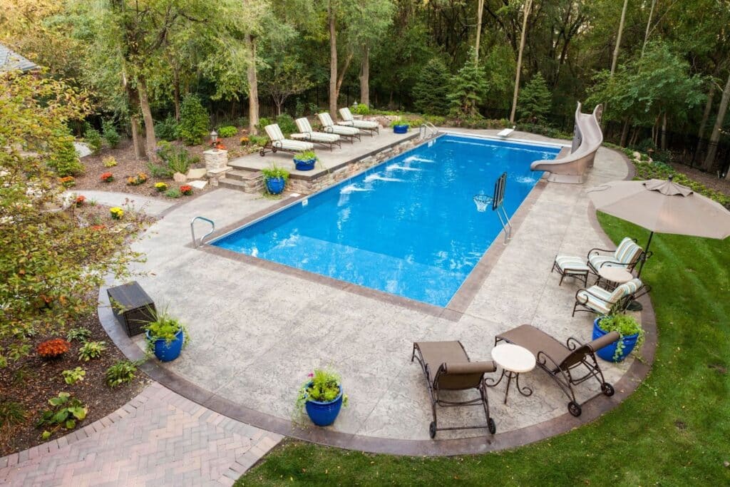 An aerial view of a backyard pool area featuring a blue swimming pool, lounge chairs, and a large water slide. Surrounding the pool are various plants in blue pots, along with a shaded seating area with chairs and a table.