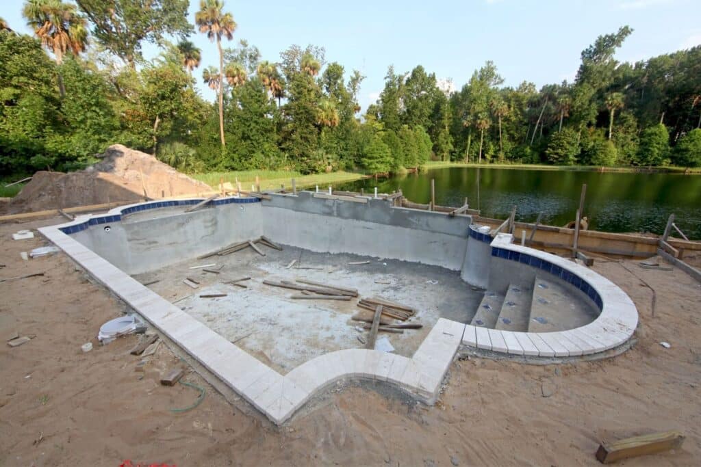 An empty, unfilled swimming pool under construction, surrounded by sand and construction materials, with a view of a lake and trees in the background.